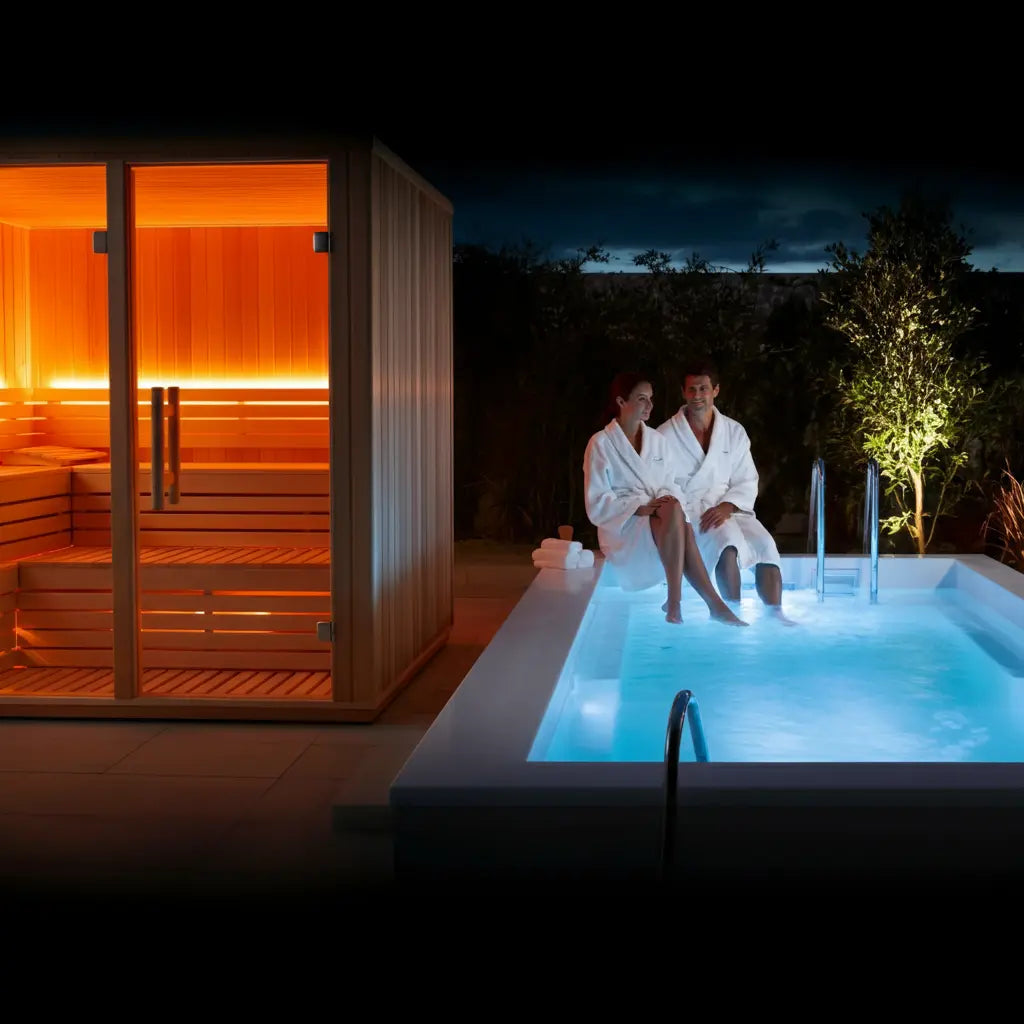 Two people in white robes sitting by a hot tub next to a wooden sauna at night.